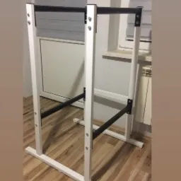 Konstrukcja metalowa, biało-czarna, prawdopodobnie do ćwiczeń, ustawiona w pomieszczeniu z drewnianą podłogą, widoczne okno z żaluzjami i grzejnik.