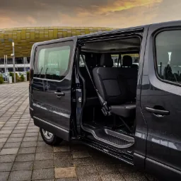 Czarny van z otwartymi drzwiami przesuwnymi, ukazujący wnętrze z czarnymi siedzeniami, na tle stadionu w Łodzi, widziany z perspektywy chodnika z kostki brukowej.