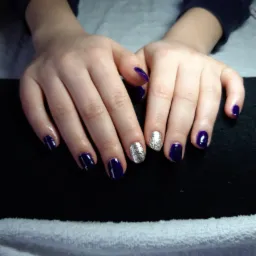 Dłonie z ciemnofioletowym lakierem na paznokciach, z akcentem brokatu na dwóch paznokciach, ułożone na białym ręczniku.