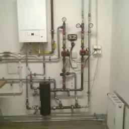 Kotłownia z białym kotłem gazowym, zbiornikiem wyrównawczym, dwoma pompami obiegowymi, manometrami i licznymi rurami w instalacji grzewczej. Widoczny również mały grzejnik panelowy oraz ekspres...