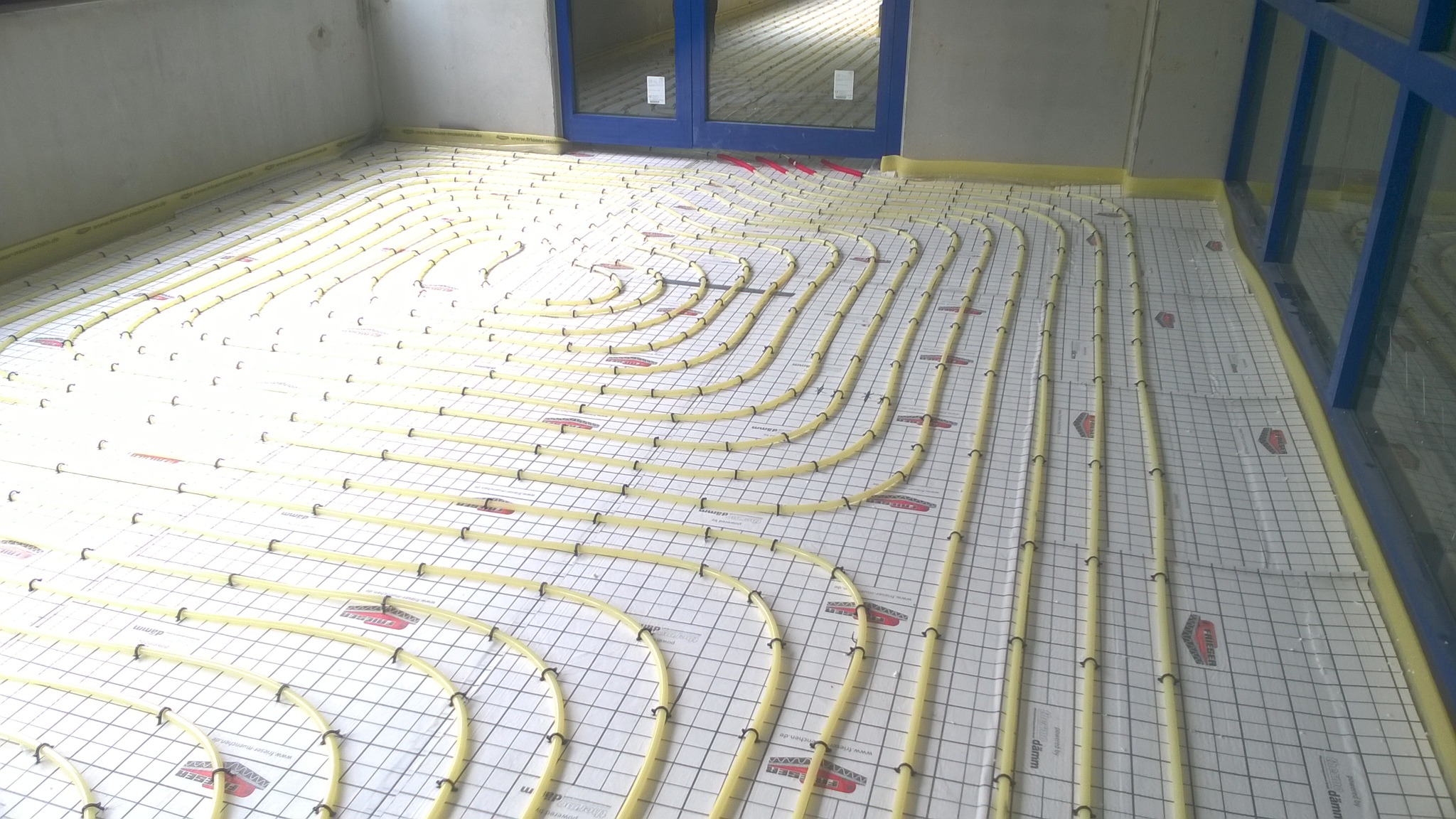 Żółte rury ogrzewania podłogowego ułożone na białej macie z nadrukowaną siatką, zabezpieczone żółtą taśmą przy ścianach i oknach.