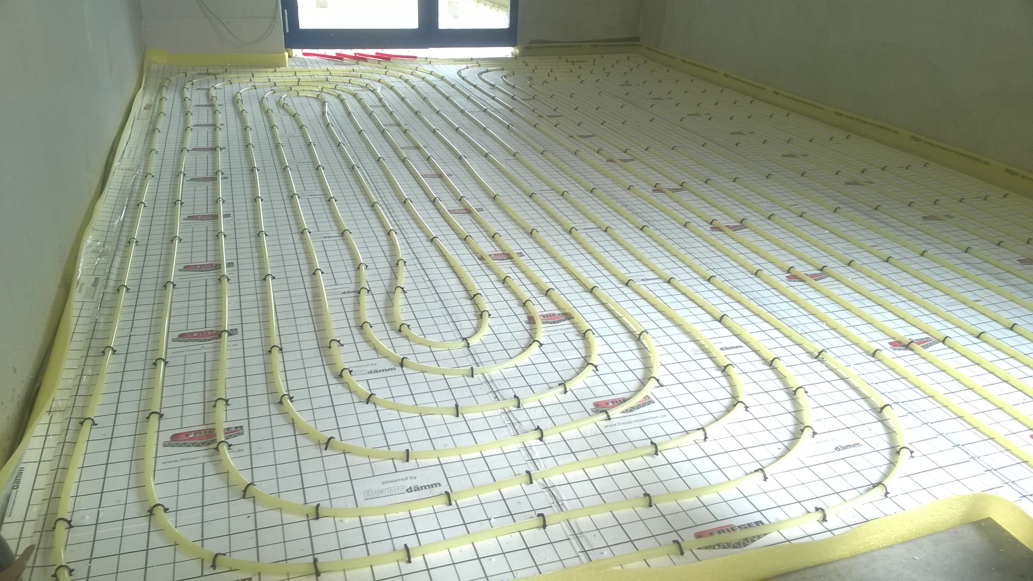 Montaż rur ogrzewania podłogowego na izolacji z siatką, widoczne mocowania i fragment okna w tle.