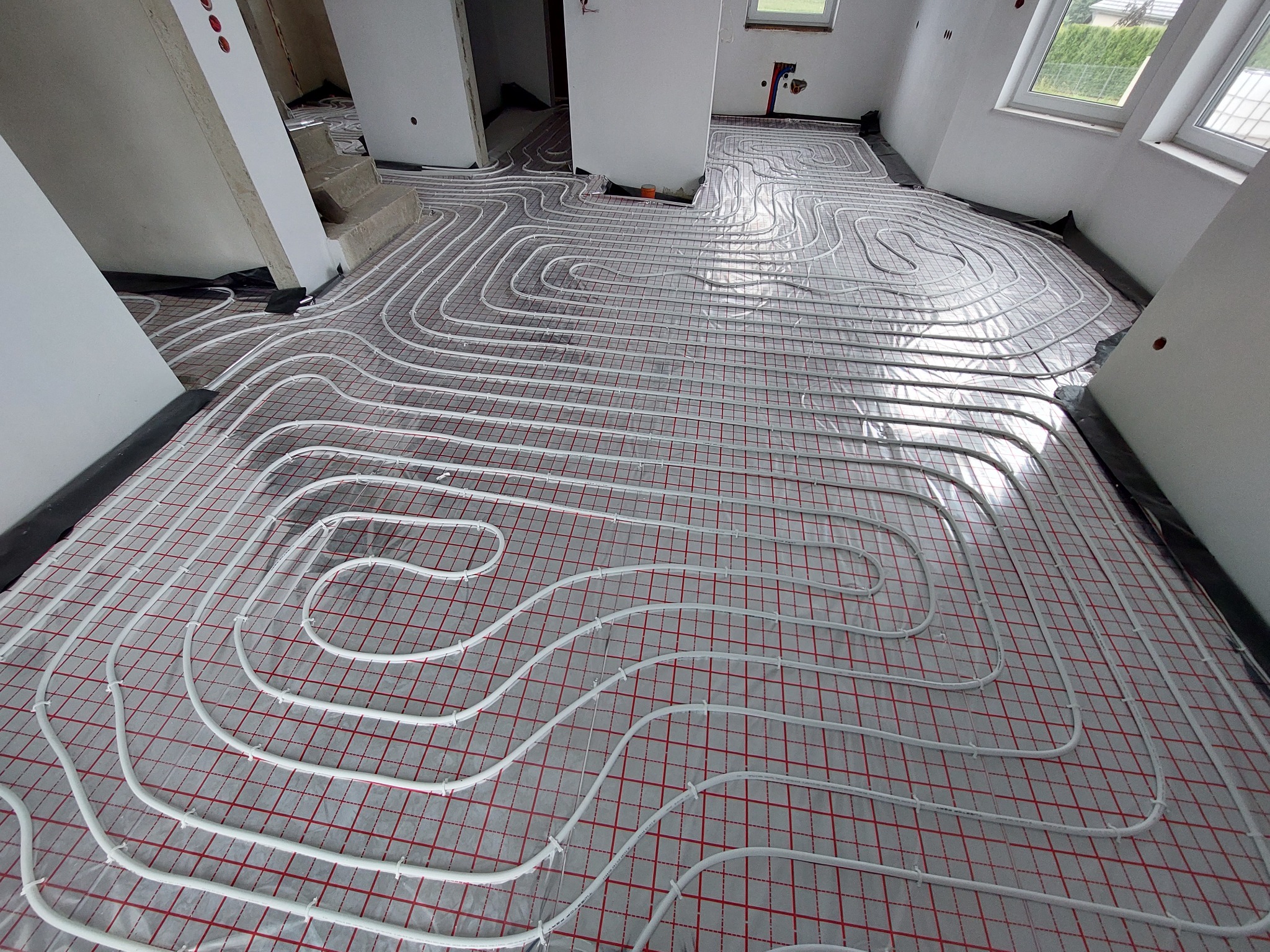 Białe rury ogrzewania podłogowego ułożone na czerwonej siatce w surowym wnętrzu, widoczne okna i przygotowane otwory w ścianie.