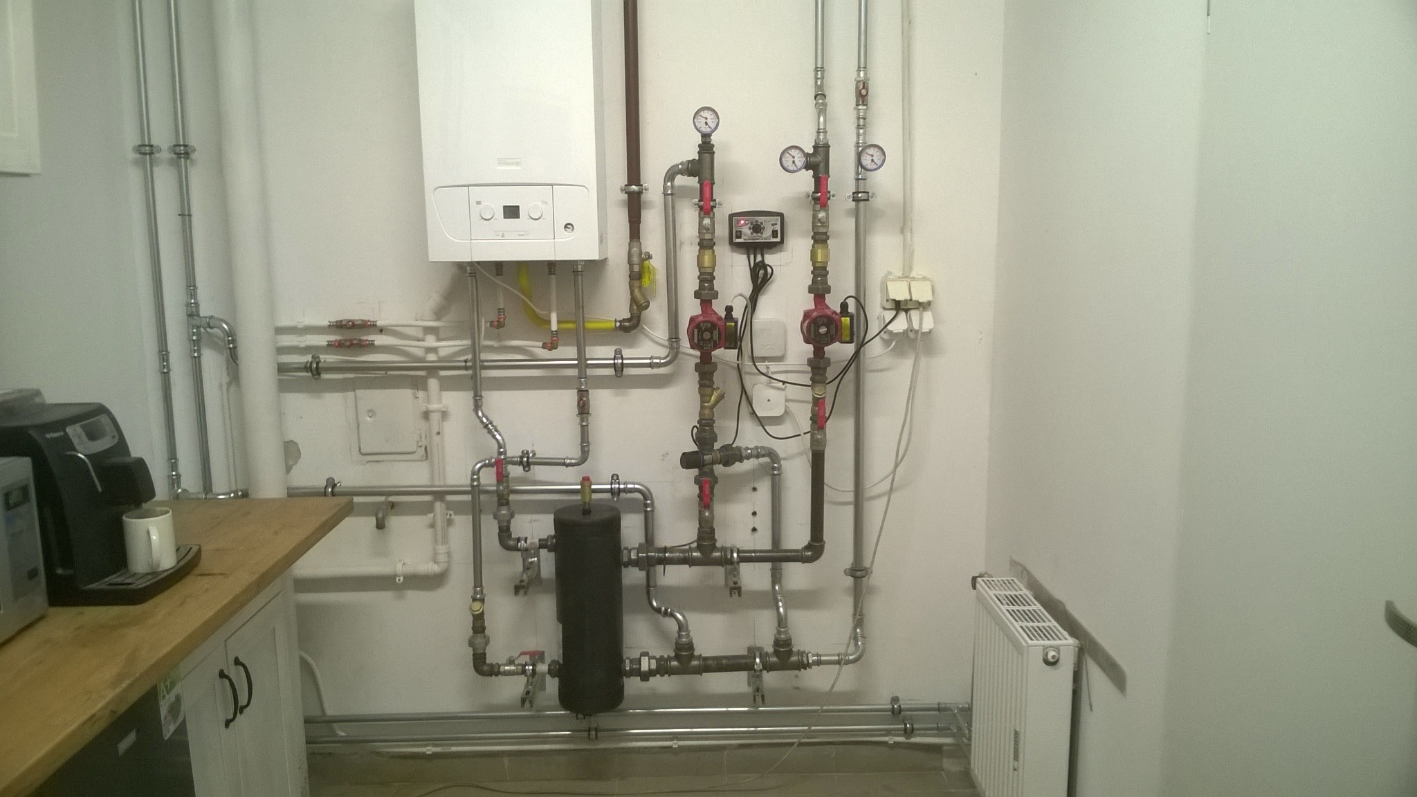Kotłownia z białym kotłem gazowym, zbiornikiem wyrównawczym, dwoma pompami obiegowymi, manometrami i licznymi rurami w instalacji grzewczej. Widoczny również mały grzejnik panelowy oraz ekspres...