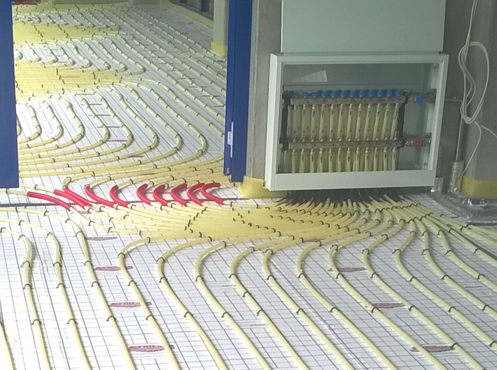 Rozłożona instalacja ogrzewania podłogowego z czerwonymi rurami na początku i rozdzielaczem w białej obudowie.