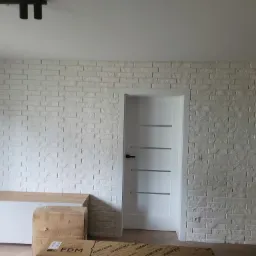 Ściana z białej cegły dekoracyjnej, oświetlenie sufitowe punktowe, biała szafka RTV i drzwi z poziomymi przeszkleniami, kartony na podłodze