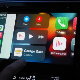 Ekran samochodowego systemu multimedialnego z interfejsem Apple CarPlay, na którym widoczna jest aplikacja do sterowania bramą garażową o statusie 'Otwarte', dotykana palcem.