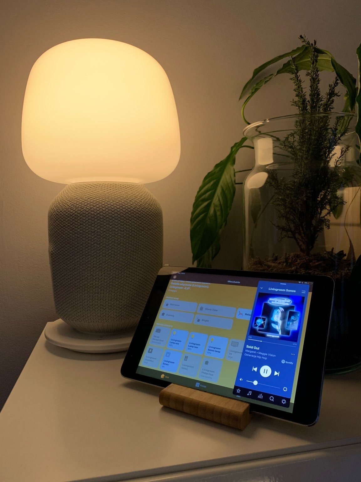 Tablet na drewnianej podstawce wyświetlający interfejs aplikacji do sterowania oświetleniem i muzyką w salonie, obok lampka w kształcie grzyba i roślina w szklanym naczyniu.