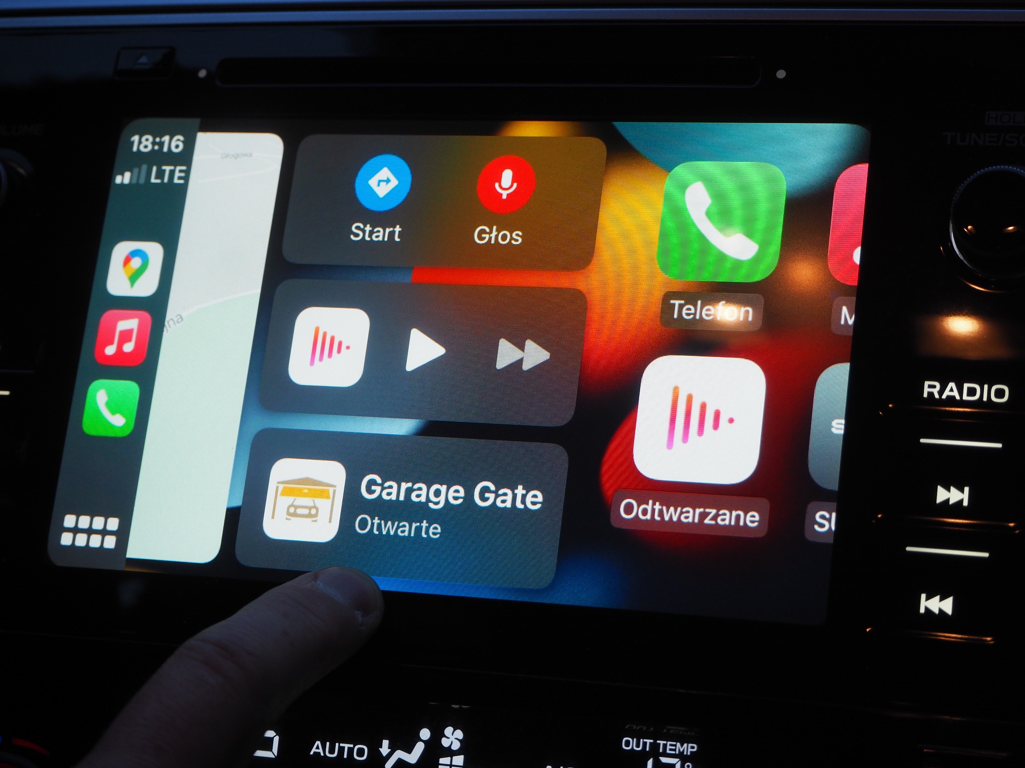 Ekran samochodowego systemu multimedialnego z interfejsem Apple CarPlay, na którym widoczna jest aplikacja do sterowania bramą garażową o statusie 'Otwarte', dotykana palcem.