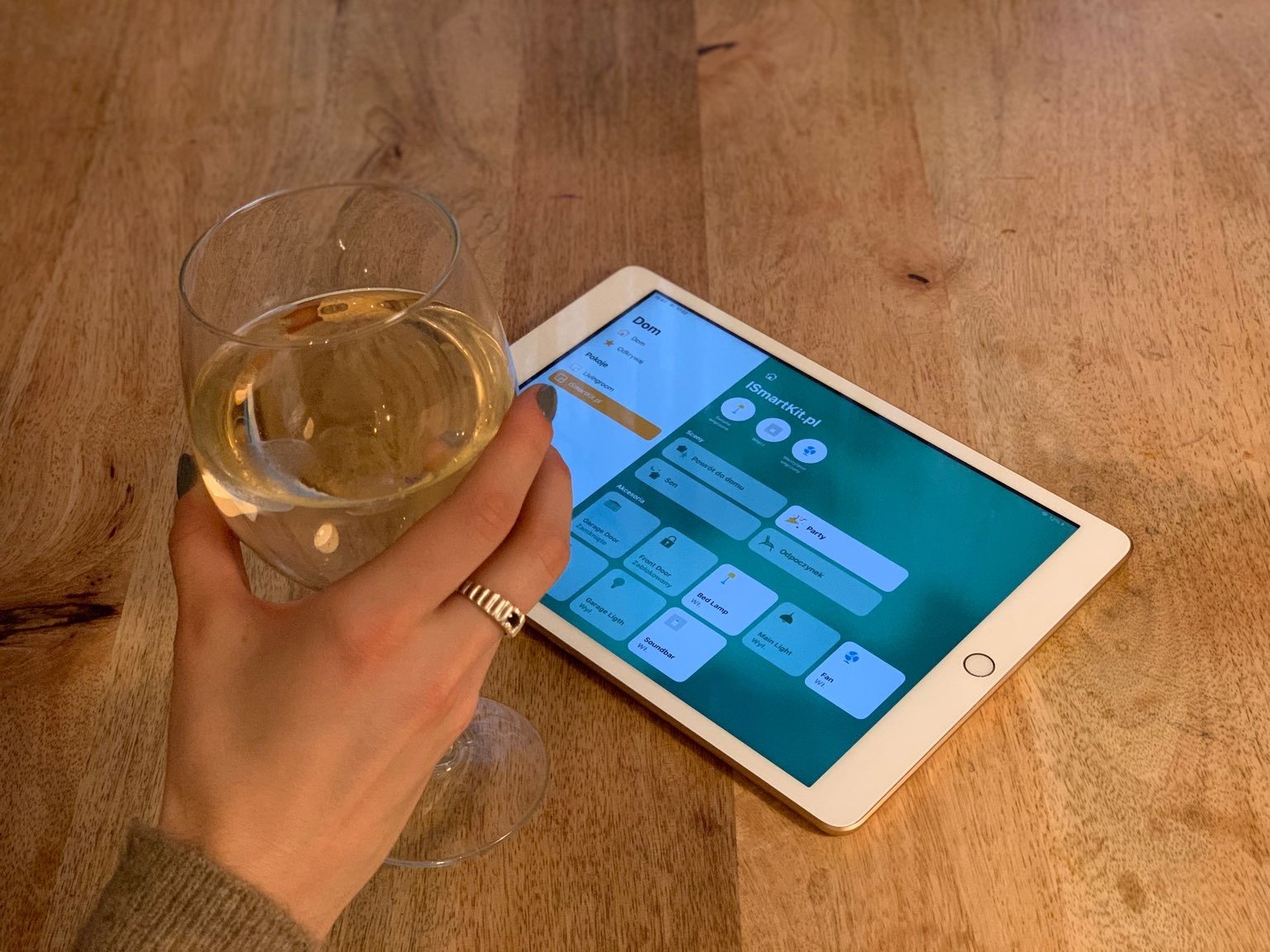 Ręka z pierścionkiem trzyma kieliszek białego wina obok tabletu z aplikacją do zarządzania domem, widok z góry na drewniany stół.