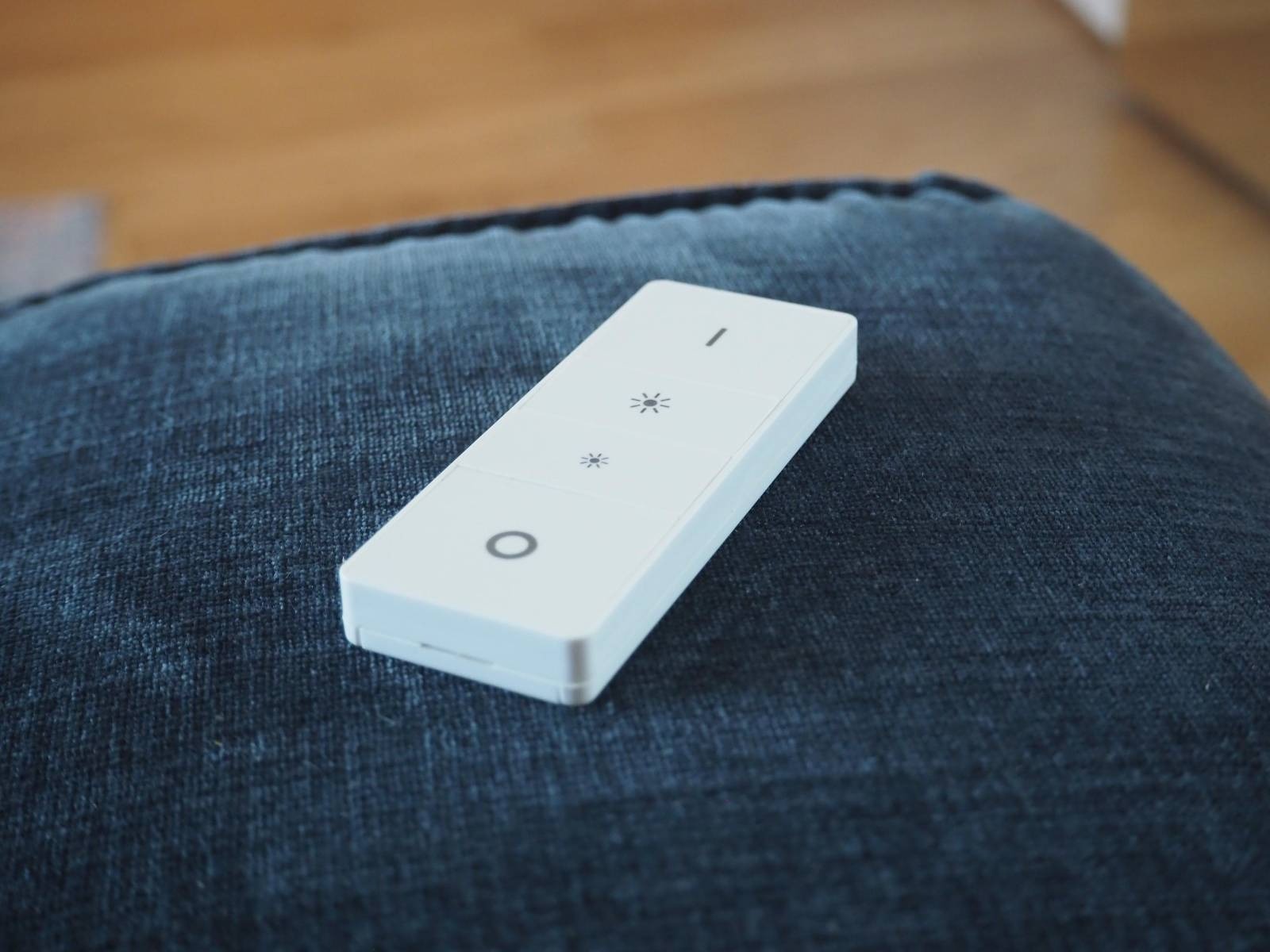 Biały pilot do sterowania oświetleniem leżący na ciemnoniebieskich jeansach, z widocznymi symbolami regulacji jasności i włącznika.