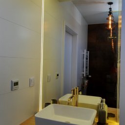 GRASTONE Emilian Stasiuk - Nowoczesna łazienka w Białymstoku z podświetlanym lustrem, białą umywalką nablatową i złotą armaturą, widoczna ściana z poziomymi panelami i dekoracyjna lampa wisząca odbijająca się w lustrze.