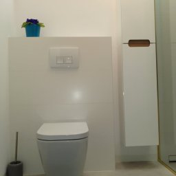 GRASTONE Emilian Stasiuk - Nowoczesna toaleta podwieszana z białym panelem zabudowy i spłuczką dwudzielną, obok wysoka szafka łazienkowa z wyfrezowanym uchwytem, na panelu doniczka z fioletowymi kwiatami, widoczna szczotka...