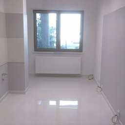 GRASTONE Emilian Stasiuk - Wnętrze pokoju po malowaniu ścian na szaro i biało, z błyszczącą białą podłogą, grzejnikiem pod oknem i widocznymi kablami wzdłuż listew przypodłogowych.