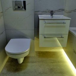 GRASTONE Emilian Stasiuk - Nowoczesna łazienka z wiszącą toaletą, szafką pod umywalką z szufladami i podświetleniem LED pod szafką, marmurowe płytki na ścianach.