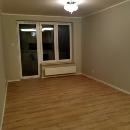 GRASTONE Emilian Stasiuk - Pusty pokój po remoncie z panelami podłogowymi w odcieniach brązu, szare ściany, białe okno z grzejnikiem pod spodem i dekoracyjna lampa sufitowa.