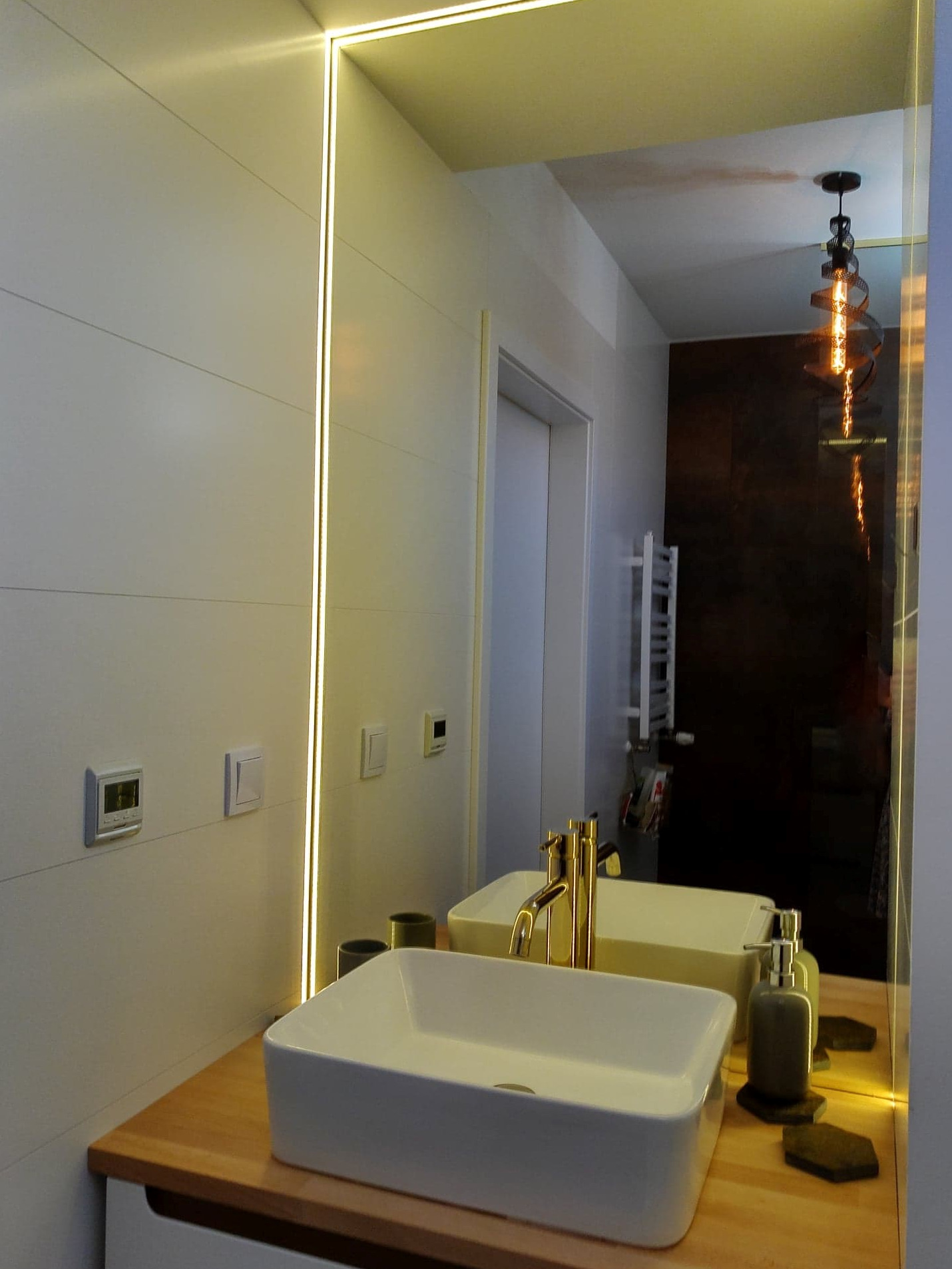 Nowoczesna łazienka w Białymstoku z podświetlanym lustrem, białą umywalką nablatową i złotą armaturą, widoczna ściana z poziomymi panelami i dekoracyjna lampa wisząca odbijająca się w lustrze.