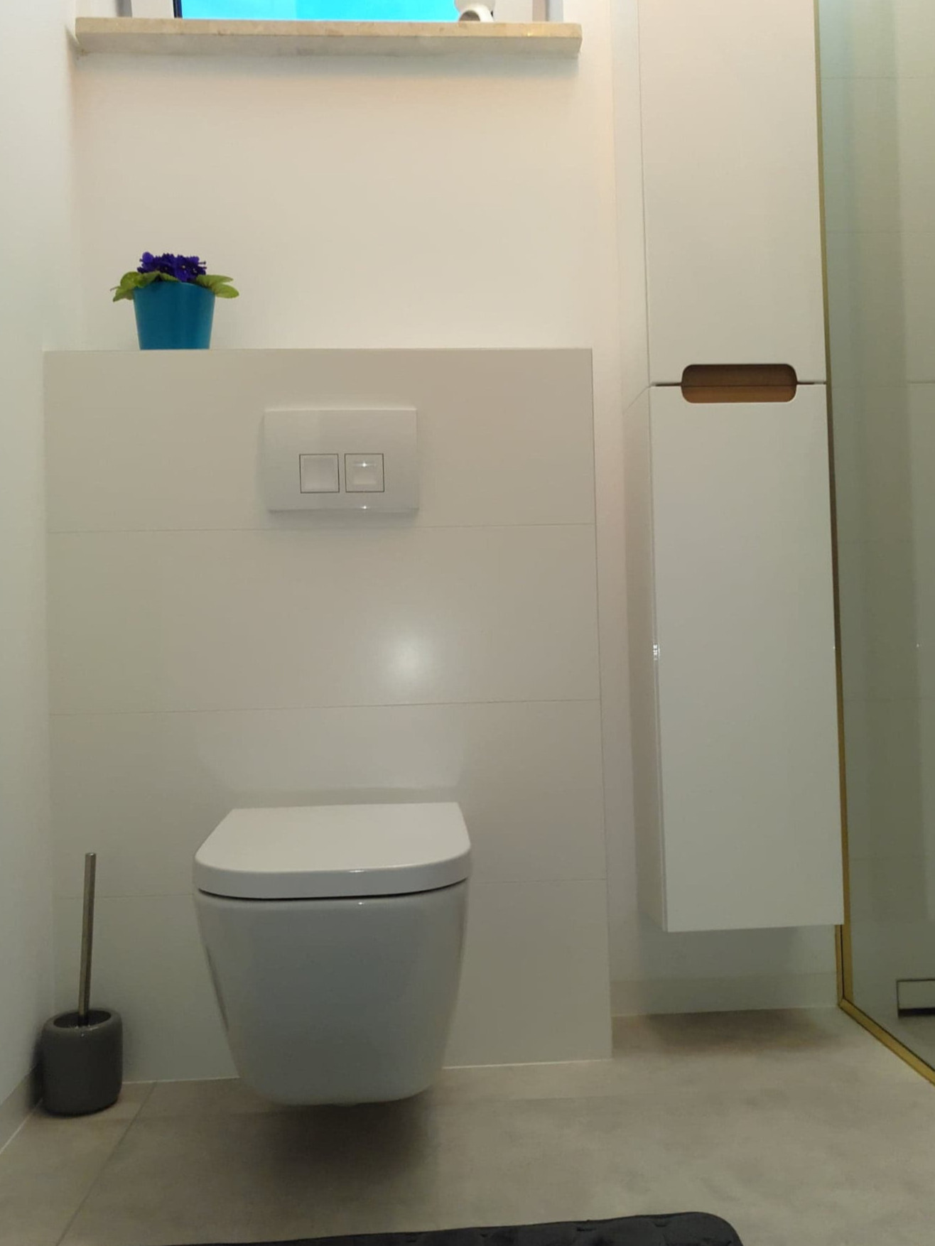 Nowoczesna toaleta podwieszana z białym panelem zabudowy i spłuczką dwudzielną, obok wysoka szafka łazienkowa z wyfrezowanym uchwytem, na panelu doniczka z fioletowymi kwiatami, widoczna szczotka...