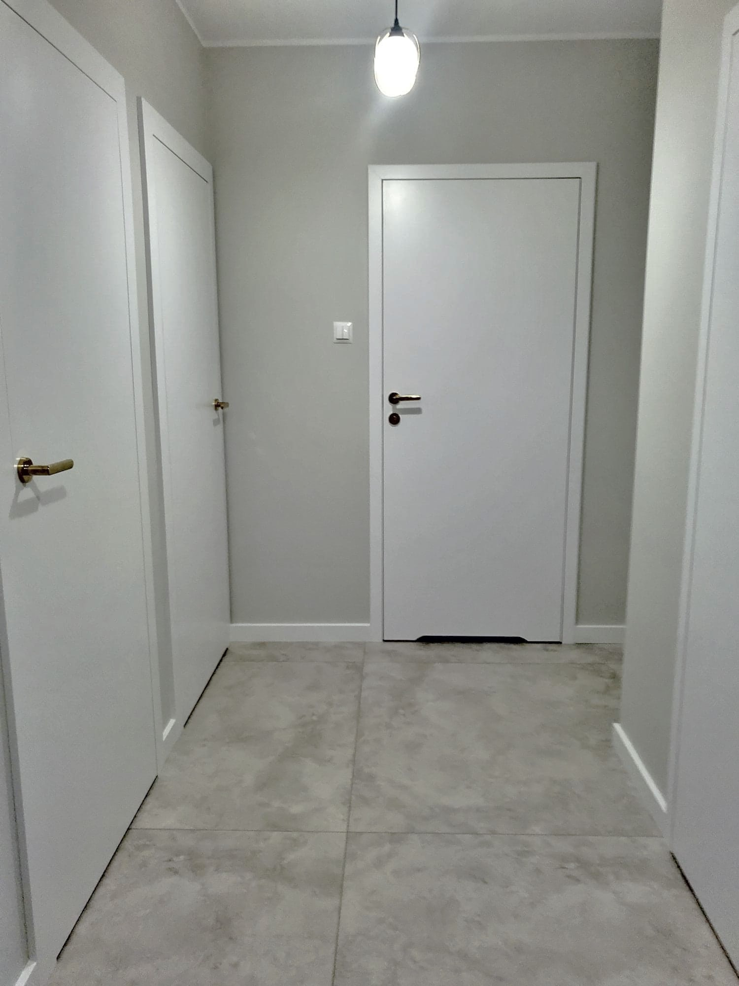 Korytarz w jasnych szarościach z trzema białymi drzwiami z mosiężnymi klamkami i szarą podłogą z płytek w formacie kwadratu, oświetlony lampą wiszącą.