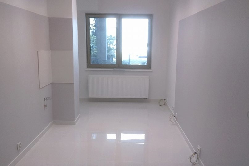 Wnętrze pokoju po malowaniu ścian na szaro i biało, z błyszczącą białą podłogą, grzejnikiem pod oknem i widocznymi kablami wzdłuż listew przypodłogowych.