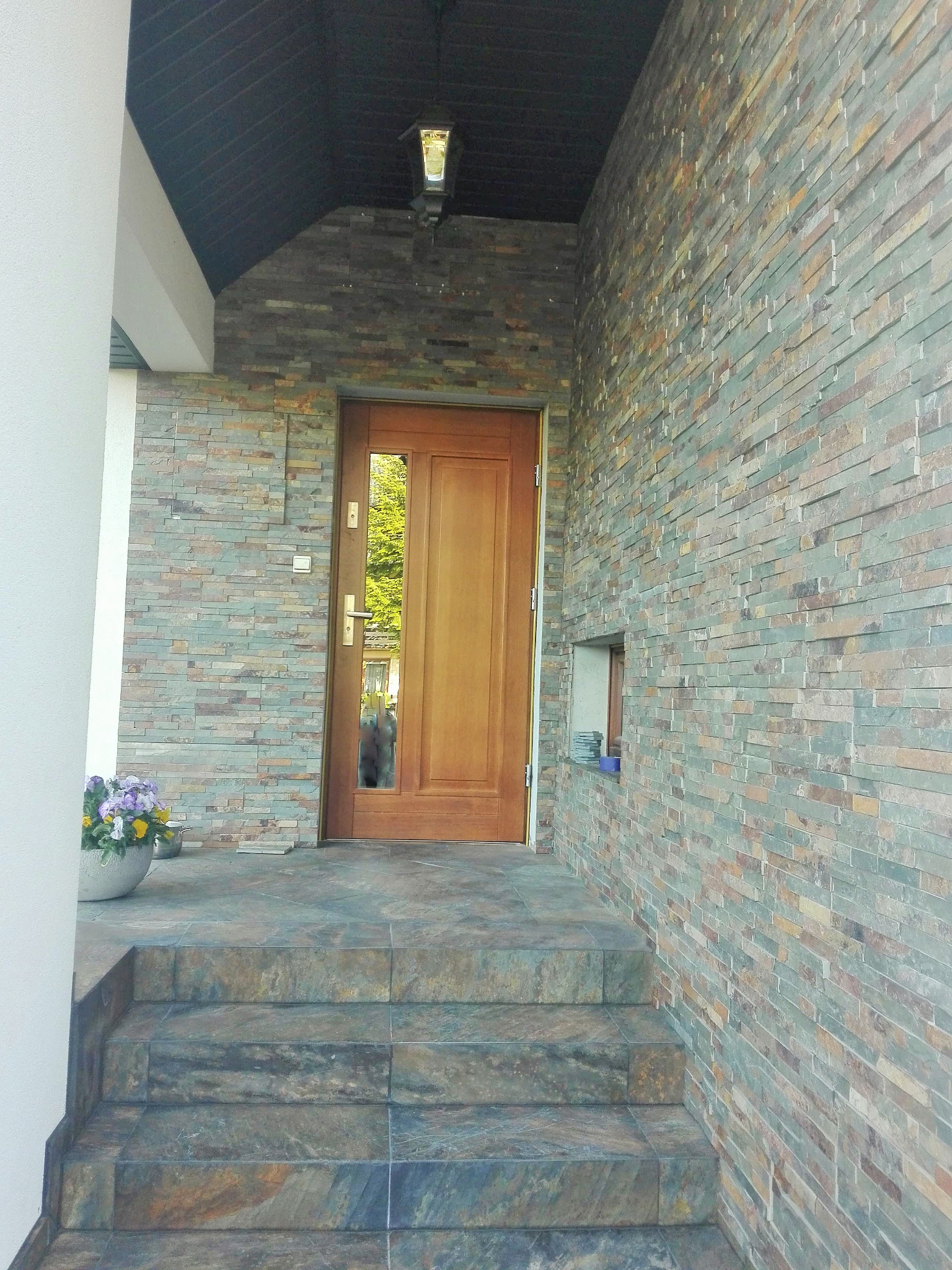 Wejście do domu z drewnianymi drzwiami, okładziną z kamienia elewacyjnego w odcieniach szarości i beżu, oraz schodami wyłożonymi płytkami imitującymi kamień. Donica z kwiatami po lewej stronie.