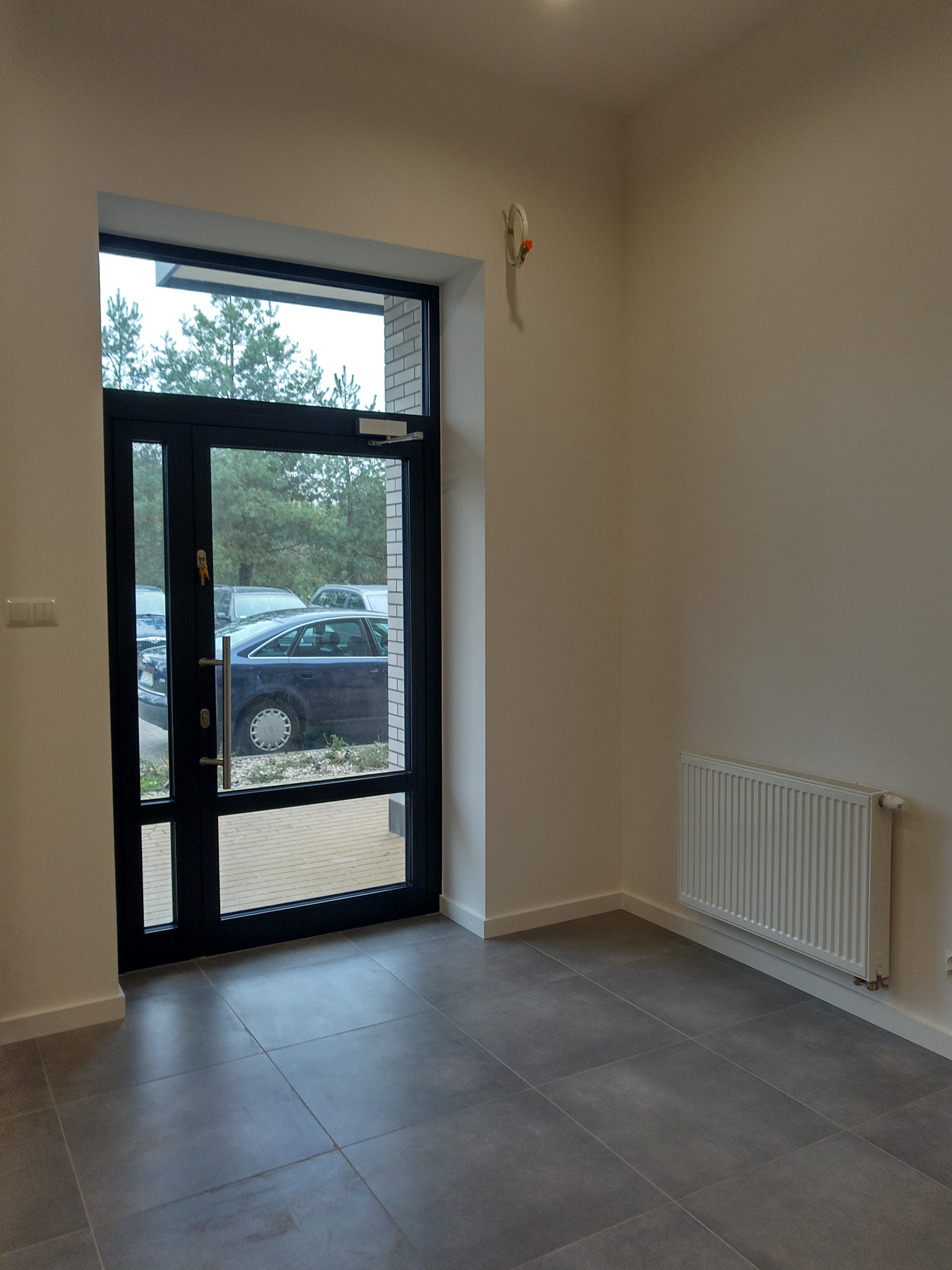 Wnętrze nowo wykończonego pomieszczenia z szarą podłogą, białymi ścianami, grzejnikiem i drzwiami balkonowymi z widokiem na parking.