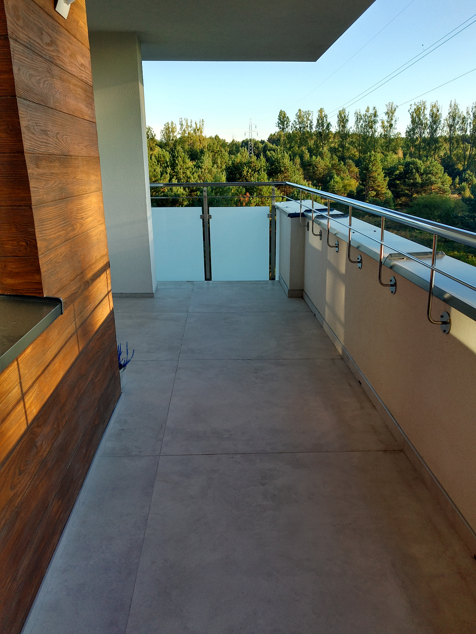 Nowoczesny balkon z szarymi płytkami, drewnianą ścianą i metalową balustradą, widok na zieleń