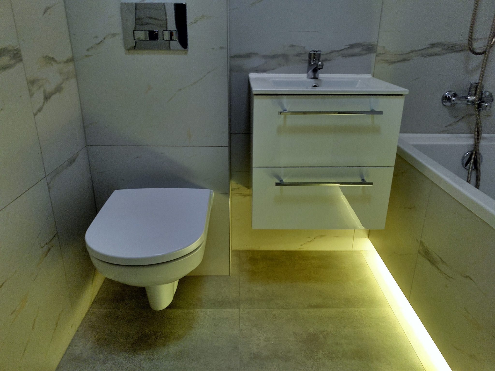 Nowoczesna łazienka z wiszącą toaletą, szafką pod umywalką z szufladami i podświetleniem LED pod szafką, marmurowe płytki na ścianach.