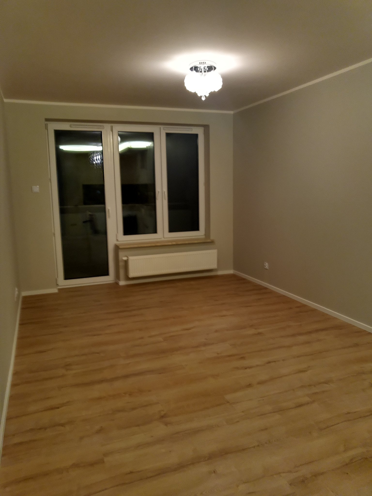 Pusty pokój po remoncie z panelami podłogowymi w odcieniach brązu, szare ściany, białe okno z grzejnikiem pod spodem i dekoracyjna lampa sufitowa.