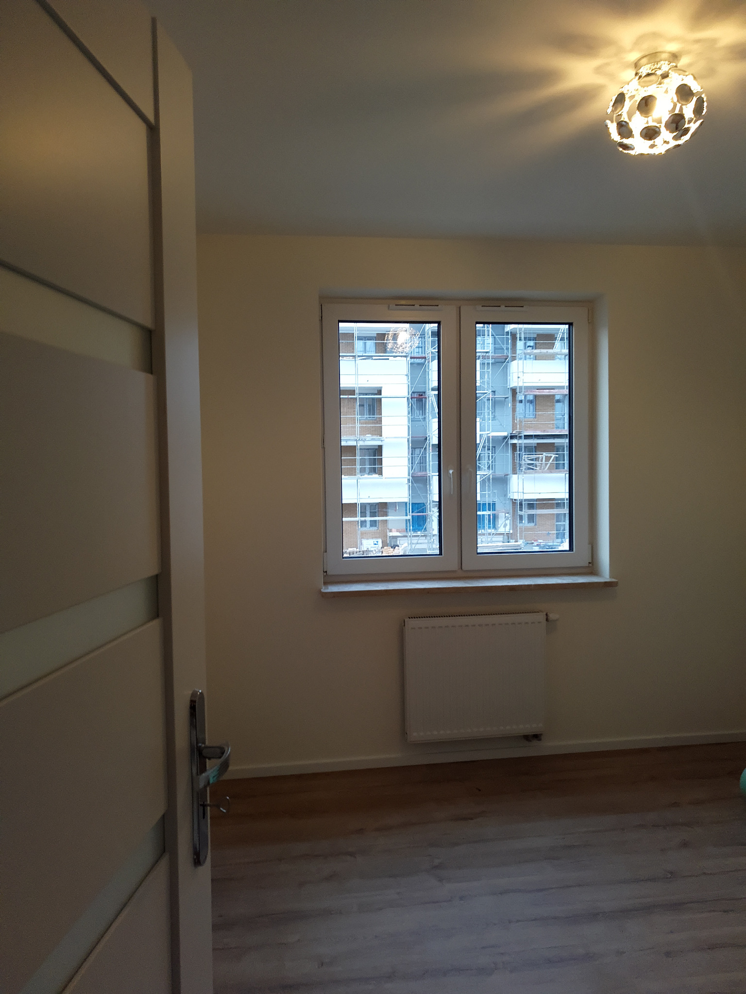 Pokój po remoncie z drewnianą podłogą, białymi ścianami, grzejnikiem pod oknem z widokiem na rusztowania budowlane za oknem i lampą sufitową w kształcie kuli.