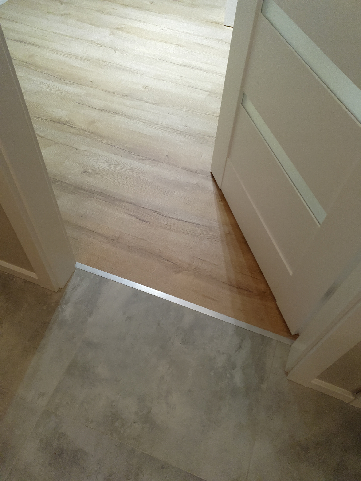 Połączenie podłogi z paneli imitujących drewno i szarych płytek ceramicznych z listwą progową, widok na otwarte drzwi wewnętrzne.