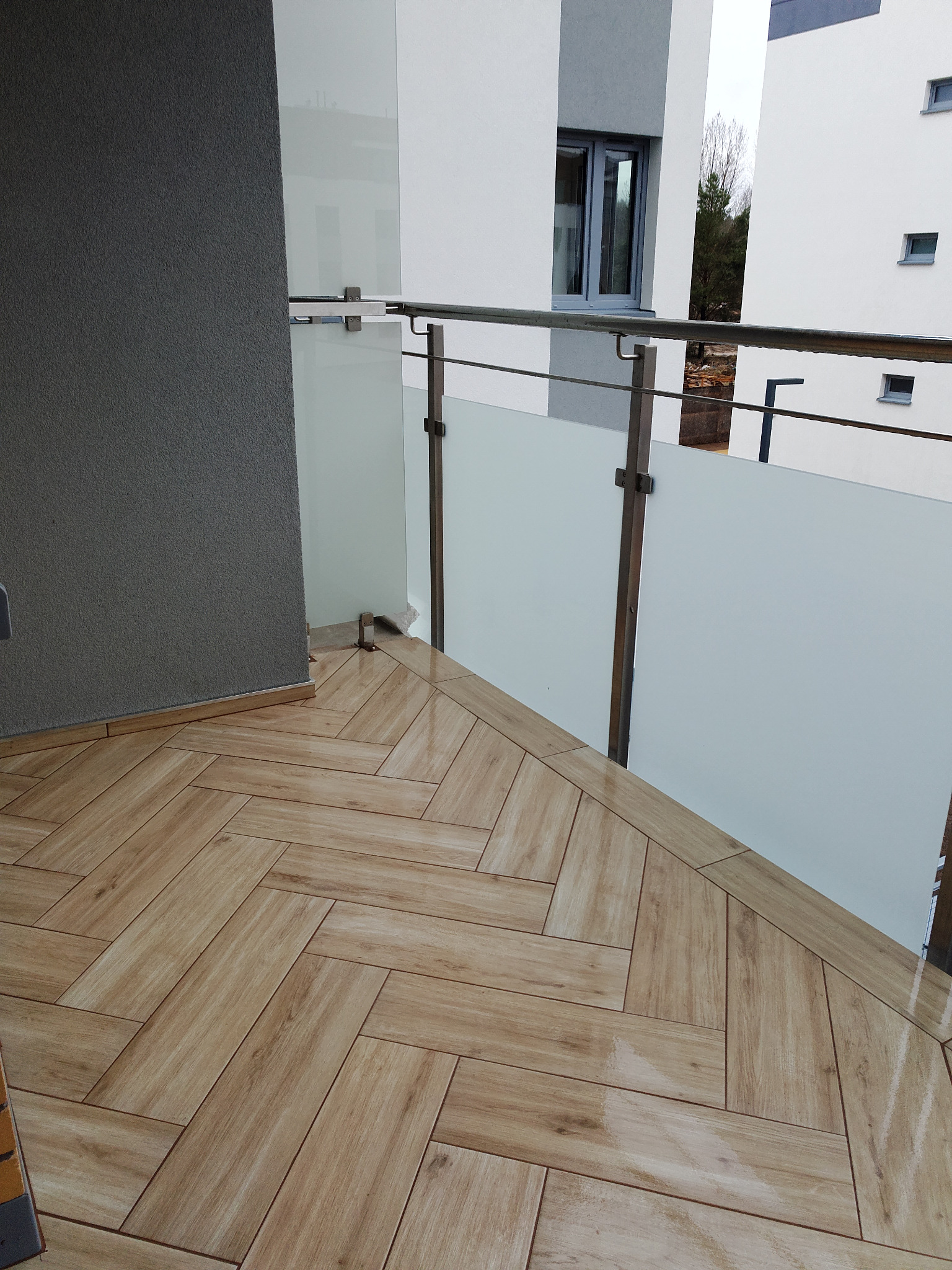 Balkon z podłogą wyłożoną płytkami imitującymi drewno ułożonymi w jodełkę, szklane balustrady z metalowymi elementami, widok na budynek mieszkalny.