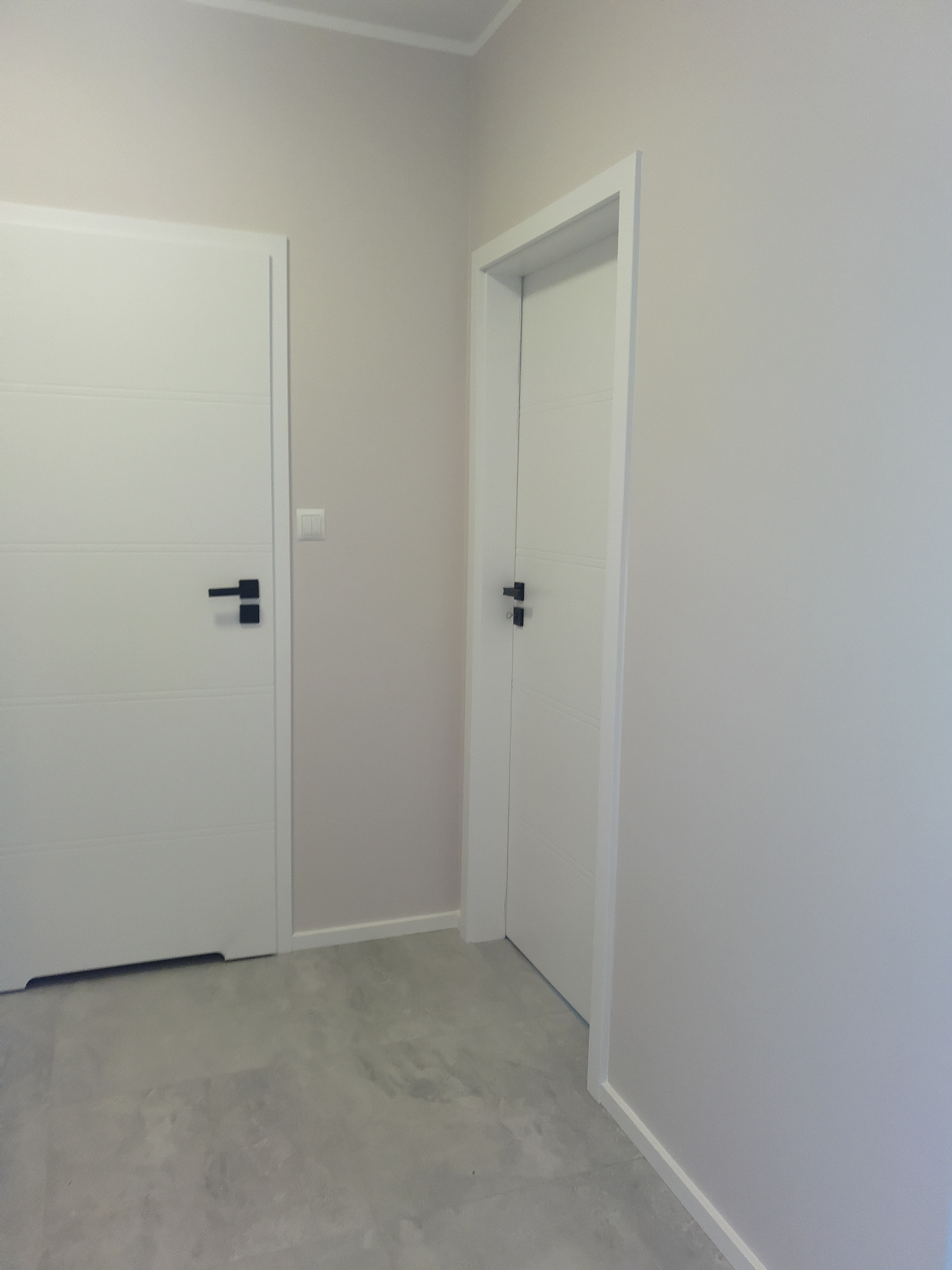 Dwa białe, nowoczesne drzwi z czarnymi klamkami w jasnym pomieszczeniu z szarą podłogą. Jedne drzwi zamknięte, drugie otwarte. Widoczny fragment listwy przypodłogowej.