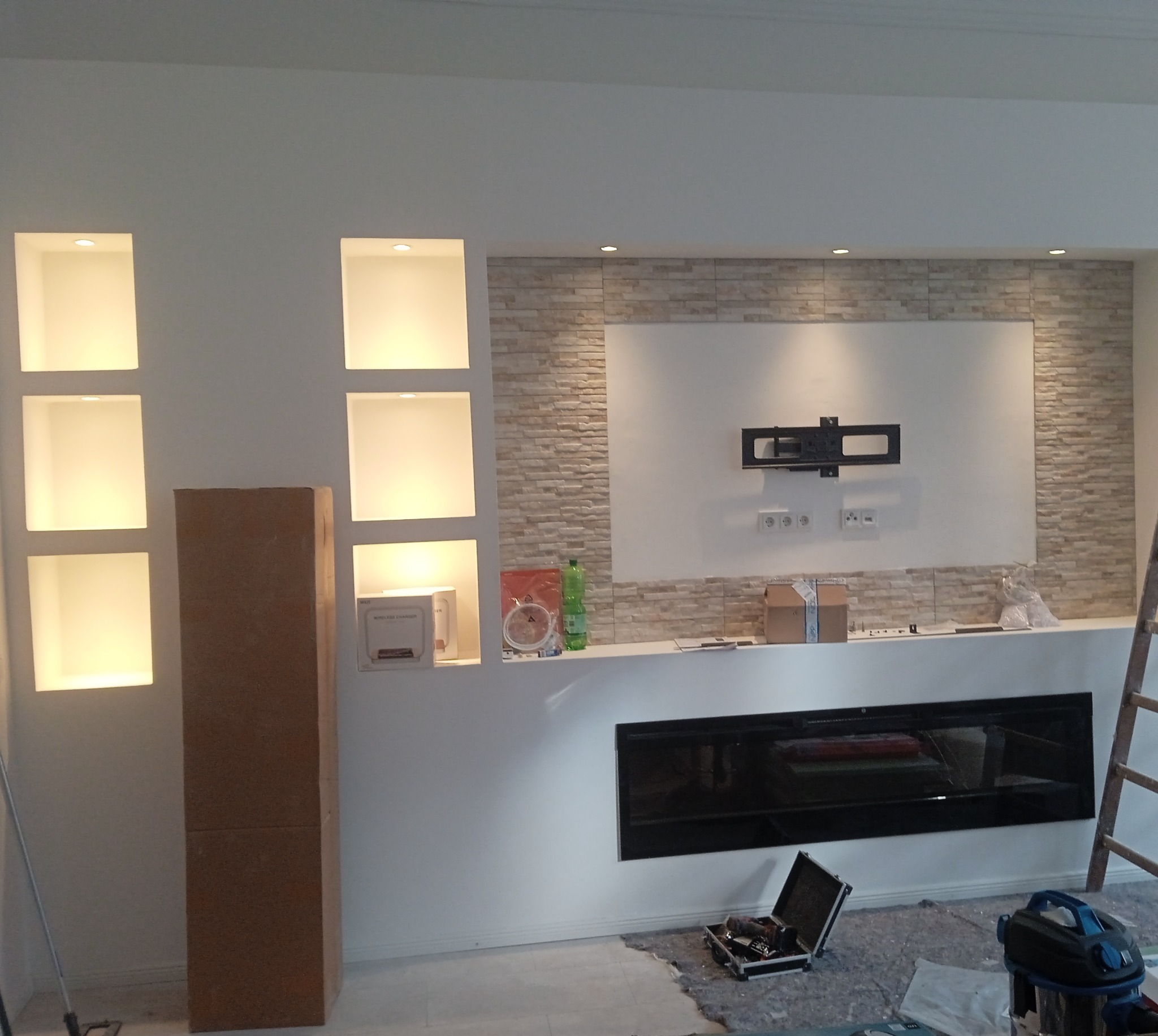 Biała ściana z wnękami oświetlonymi punktowo, dekoracyjna ściana z imitacji kamienia, zamontowany uchwyt na telewizor i czarny wkład kominkowy, widoczne narzędzia i akcesoria remontowe.