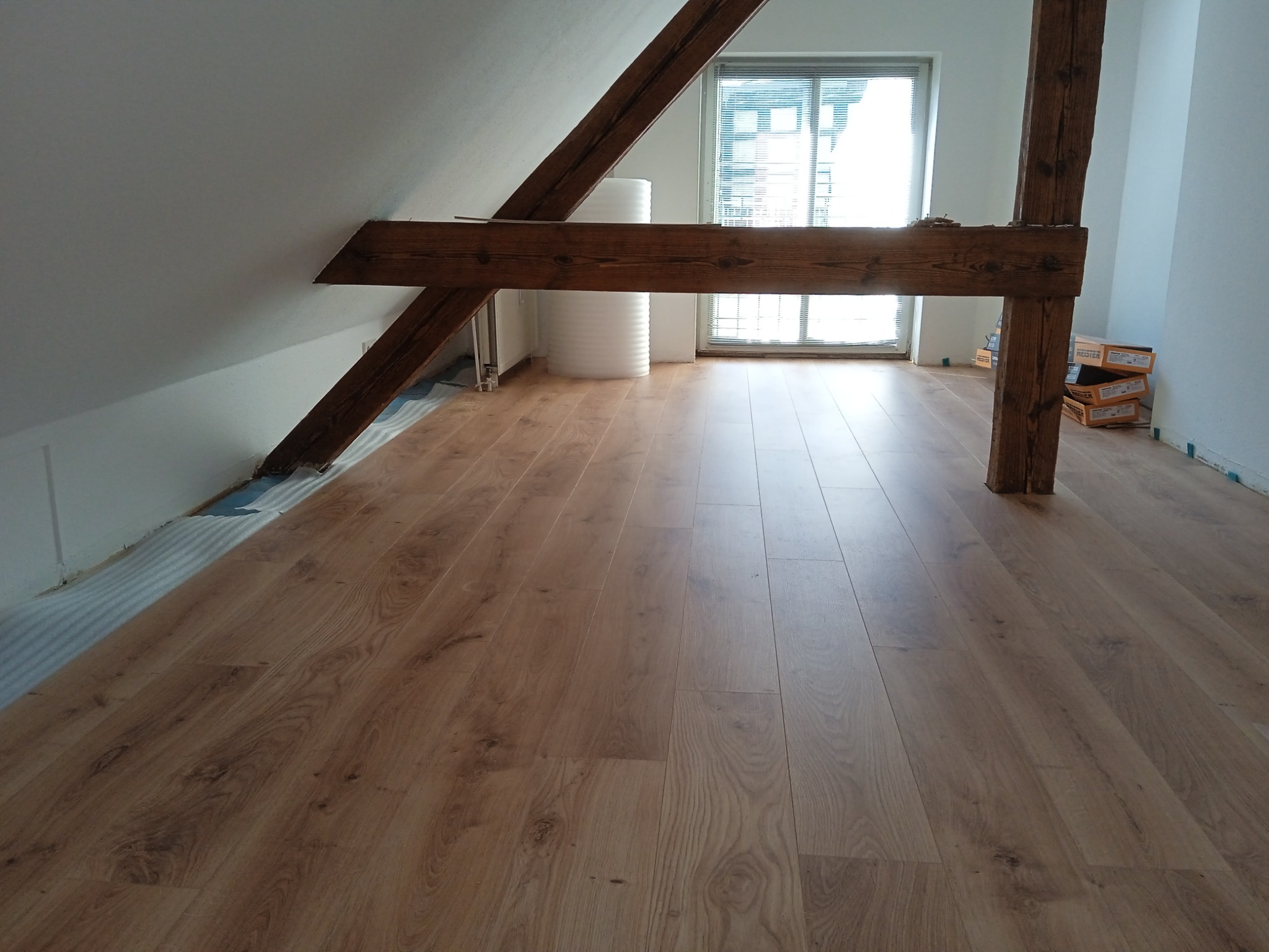Ukończona podłoga z paneli w pokoju na poddaszu z drewnianymi belkami w Grodzisku Wielkopolskim, widoczne rolki folii izolacyjnej i resztki opakowań po panelach.
