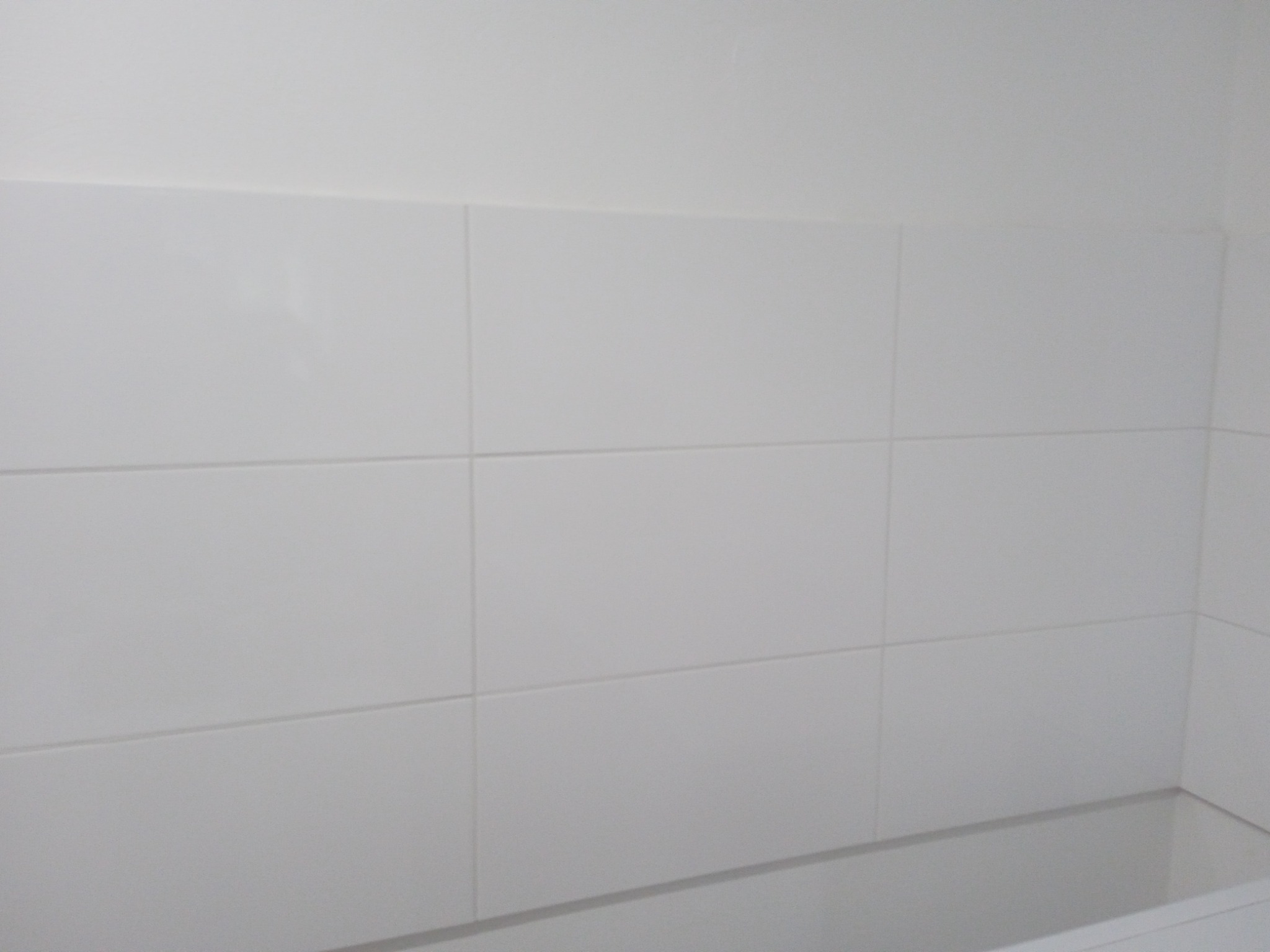 Białe, prostokątne płytki ceramiczne na ścianie nad wanną, widoczne fugi i krawędź wanny.