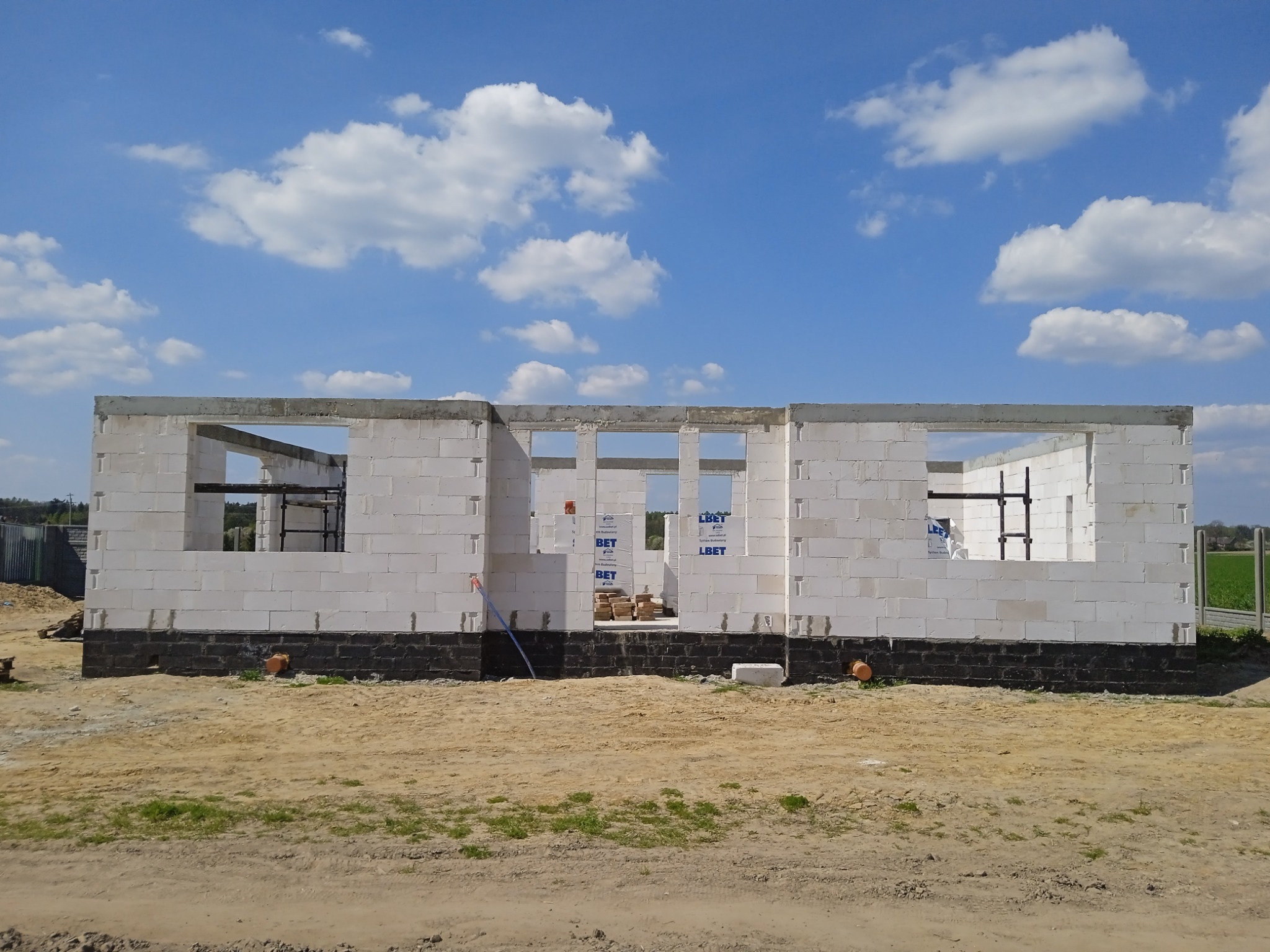 Surowy stan budynku jednorodzinnego z białych bloczków, z widoczną czarną izolacją fundamentów, na tle błękitnego nieba z chmurami.