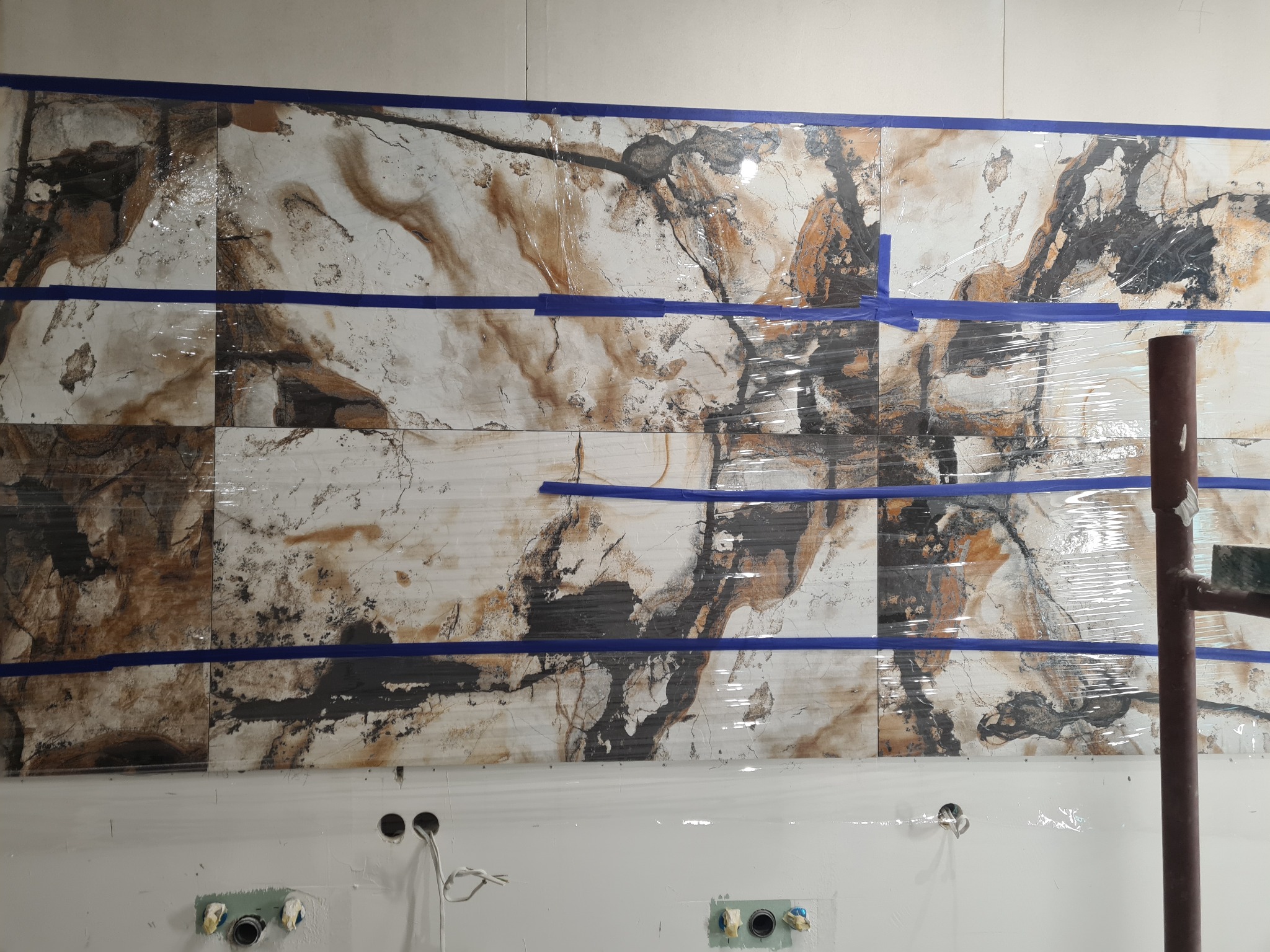 Montaż płytek imitujących marmur na ścianie, zabezpieczonych folią i niebieską taśmą malarską, widoczne przygotowane przyłącza wodno-kanalizacyjne.