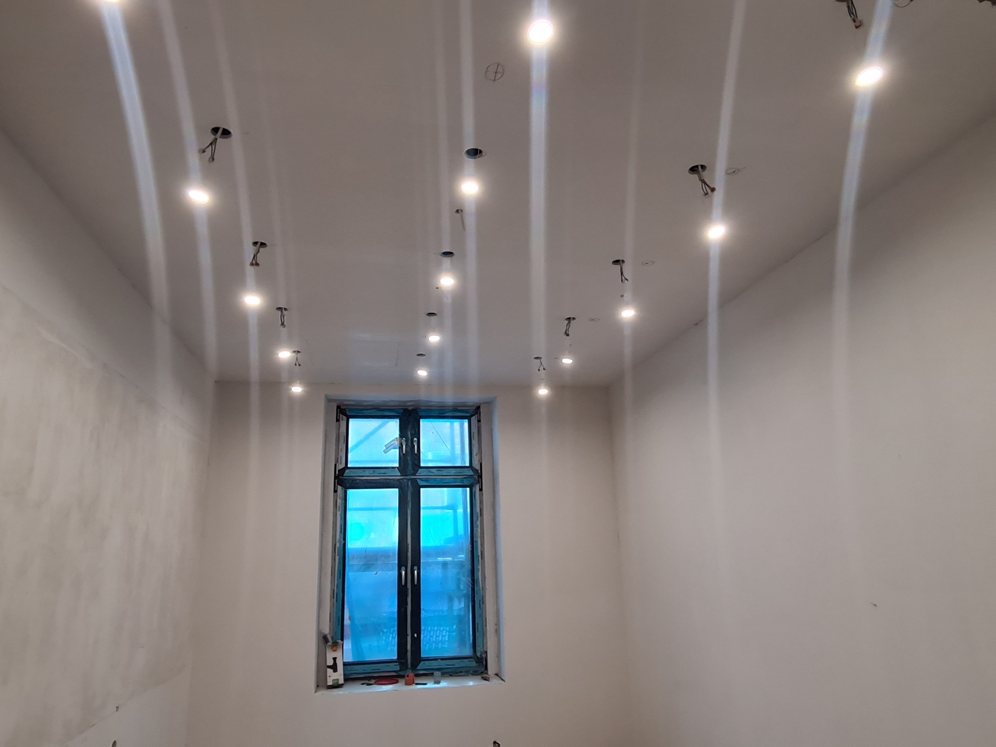 Wnętrze kamienicy z widocznymi punktami świetlnymi w suficie podwieszanym oraz oknem zabezpieczonym folią malarską.