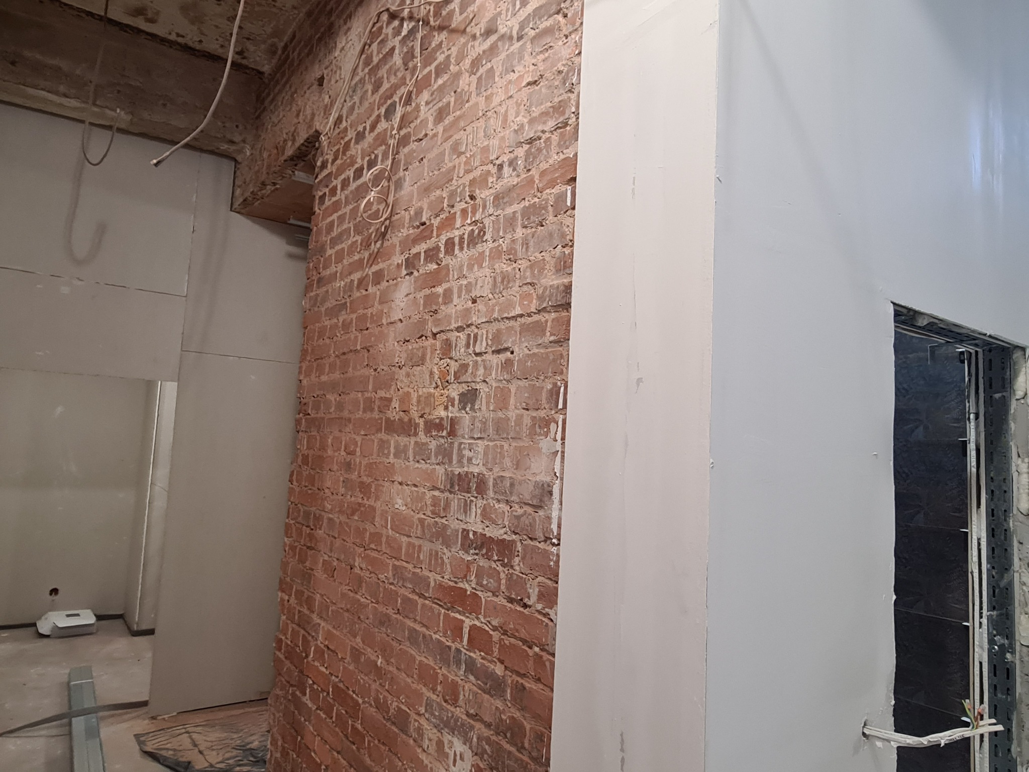 Wnętrze w trakcie prac wykończeniowych: fragment ściany z surowej czerwonej cegły sąsiaduje z fragmentem ściany pomalowanej na szaro, widoczne przewody elektryczne i surowy sufit.