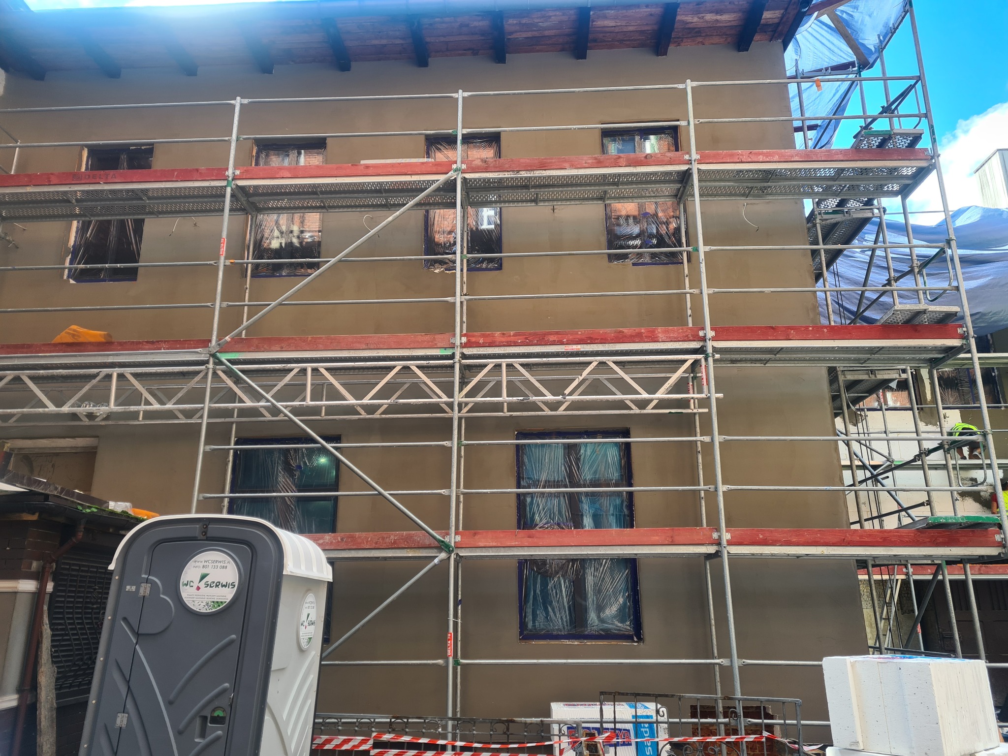 Kamienica w Tarnowskich Górach w trakcie prac elewacyjnych: świeżo otynkowana ściana z zakrytymi oknami na rusztowaniu, widoczna przenośna toaleta i materiały budowlane.