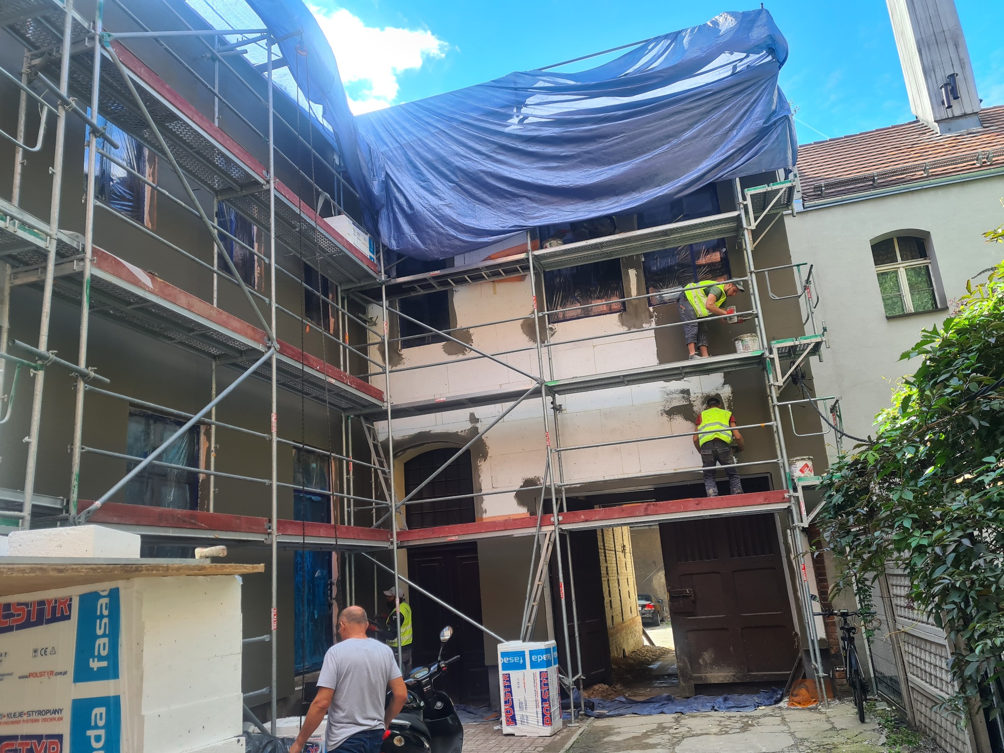 Ocieplanie ścian kamienicy z lokalami usługowymi: robotnicy na rusztowaniu aplikują warstwę izolacyjną, zabezpieczenie plandeką, widoczne paczki styropianu.