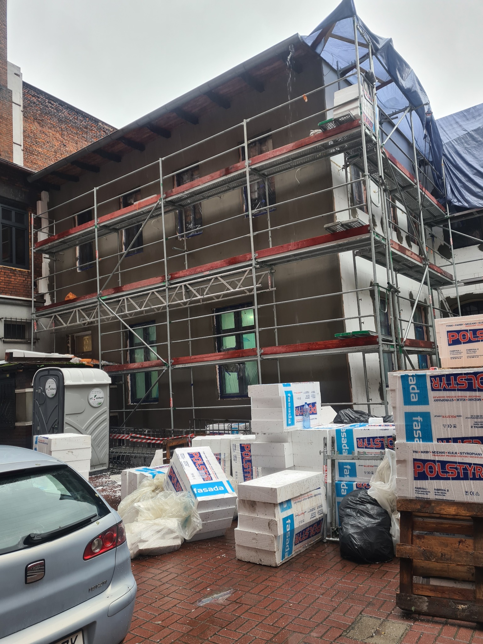 Kamienica z lokalami usługowymi w trakcie ocieplania, widoczne rusztowania, styropian fasadowy Polstyr, przenośna toaleta i zaparkowany samochód osobowy.