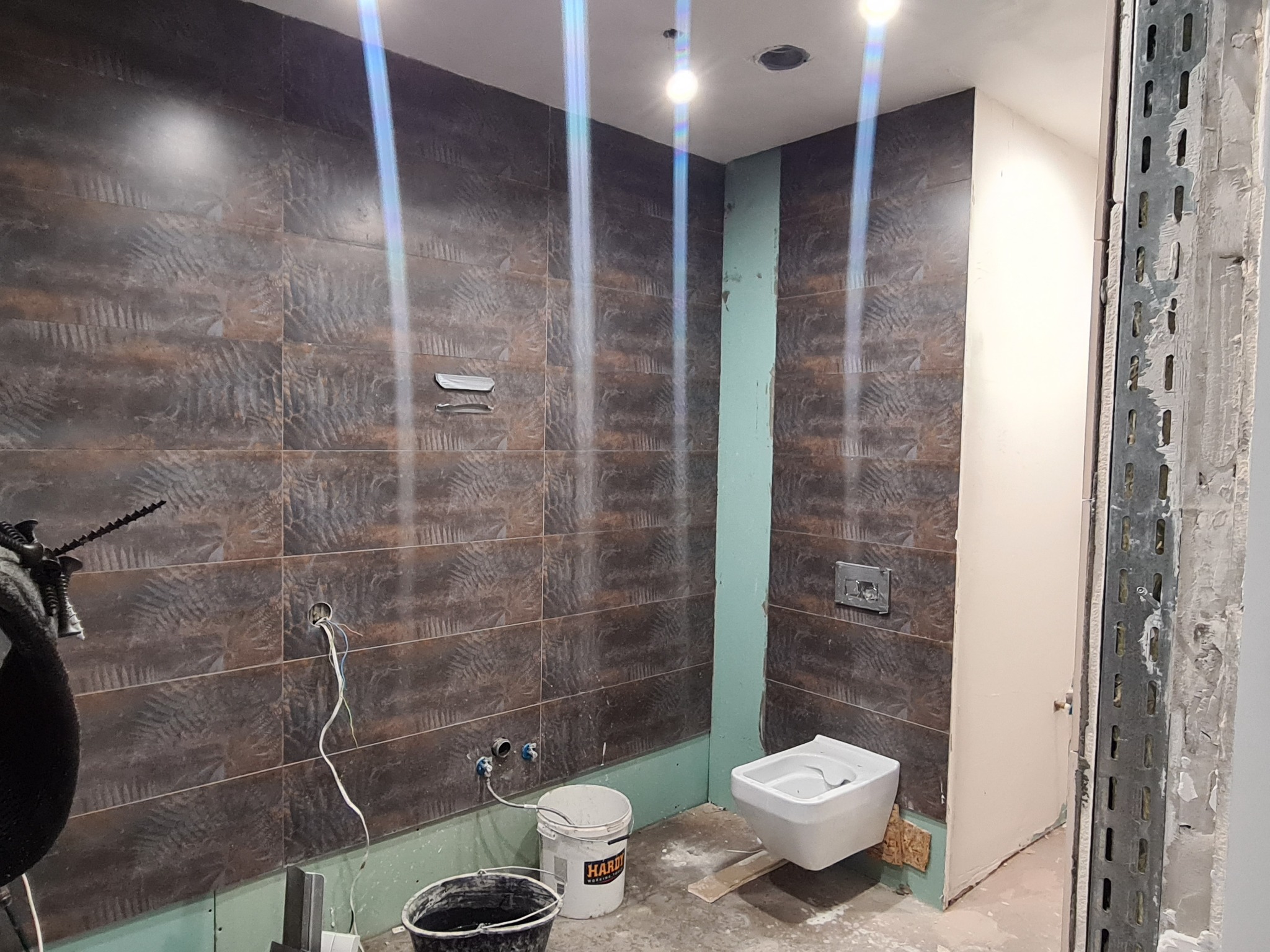 Wnętrze łazienki w trakcie prac wykończeniowych, widoczne brązowe płytki z wzorem na ścianach, biały bidet, wiadro z zaprawą, wystające przewody elektryczne i fragment konstrukcji nośnej przy wejściu.