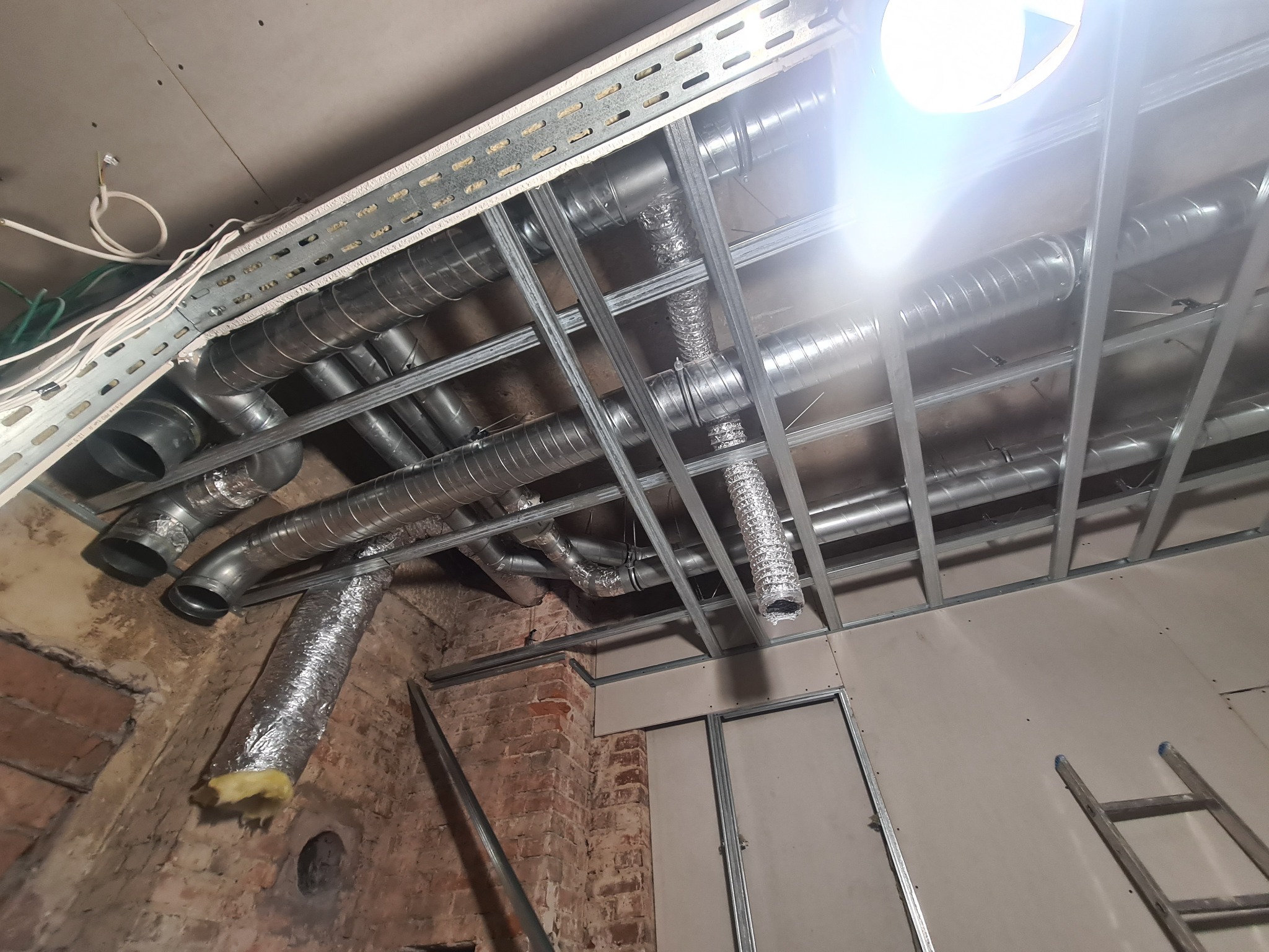 Montaż instalacji wentylacyjnej pod sufitem podwieszanym w kamienicy z odsłoniętą cegłą, widoczne rury wentylacyjne aluminiowe i stalowe, konstrukcja metalowa sufitu oraz fragment ściany z cegły.