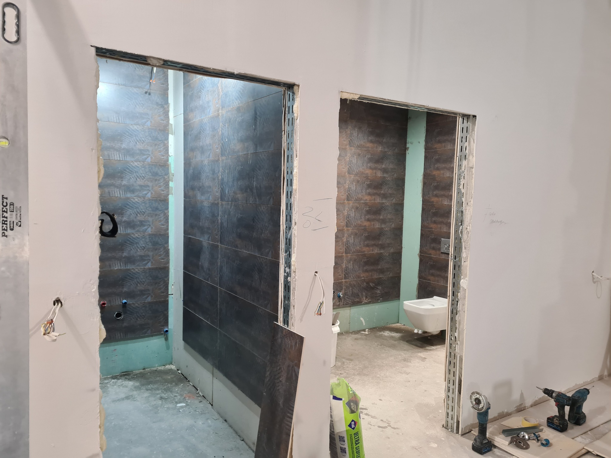 Dwa widoki na łazienkę w trakcie remontu: w jednym wykończony prysznic z ciemnymi płytkami, w drugim częściowo wykończona toaleta z wiszącą muszlą, widoczne narzędzia i materiały budowlane.