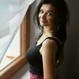 Kobieta z ciemnymi włosami, ubrana w czarny top i kwiecistą spódnicę, stoi przy oknie z rozwianą białą firanką, spoglądając z uśmiechem przez ramię.