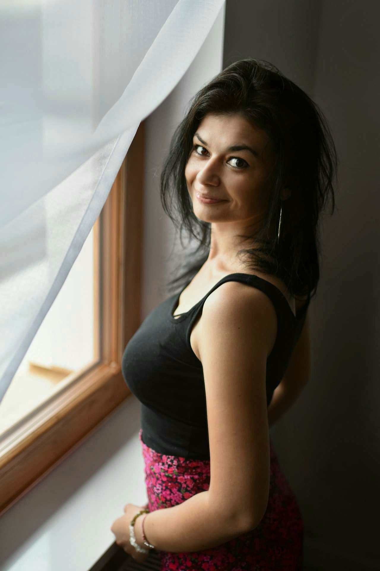 Kobieta z ciemnymi włosami, ubrana w czarny top i kwiecistą spódnicę, stoi przy oknie z rozwianą białą firanką, spoglądając z uśmiechem przez ramię.