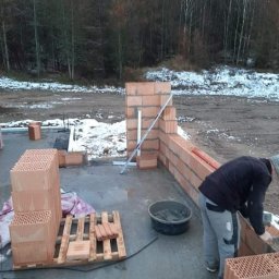 Michał Leszkowski - Wznoszenie ściany z bloczków ceramicznych Porotherm na fundamencie w zimowej scenerii, widoczna zaprawa w wiadrze i paleta z bloczkami.