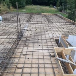 Michał Leszkowski - Zbrojenie płyty fundamentowej z drewnianym szalunkiem i wystającymi prętami pionowymi na tle zieleni.